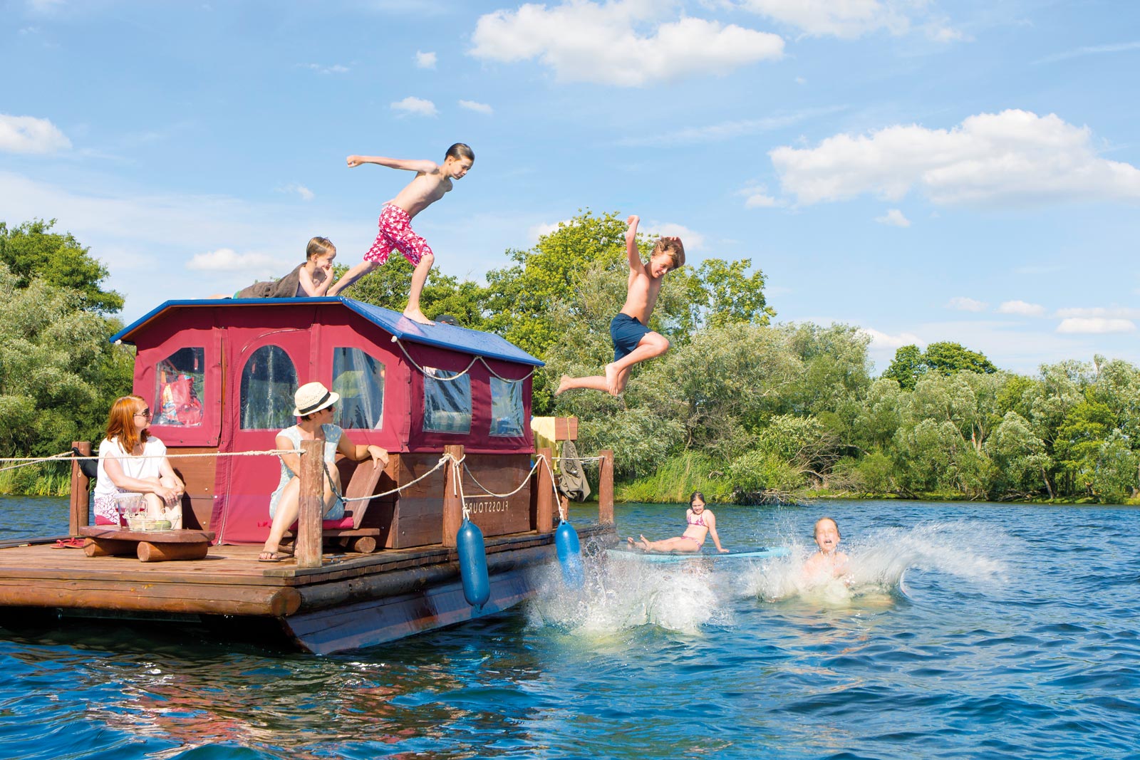 Familie mit Kindern auf Floß, die dabei ins Wasser springen, bei sonnigem Wetter